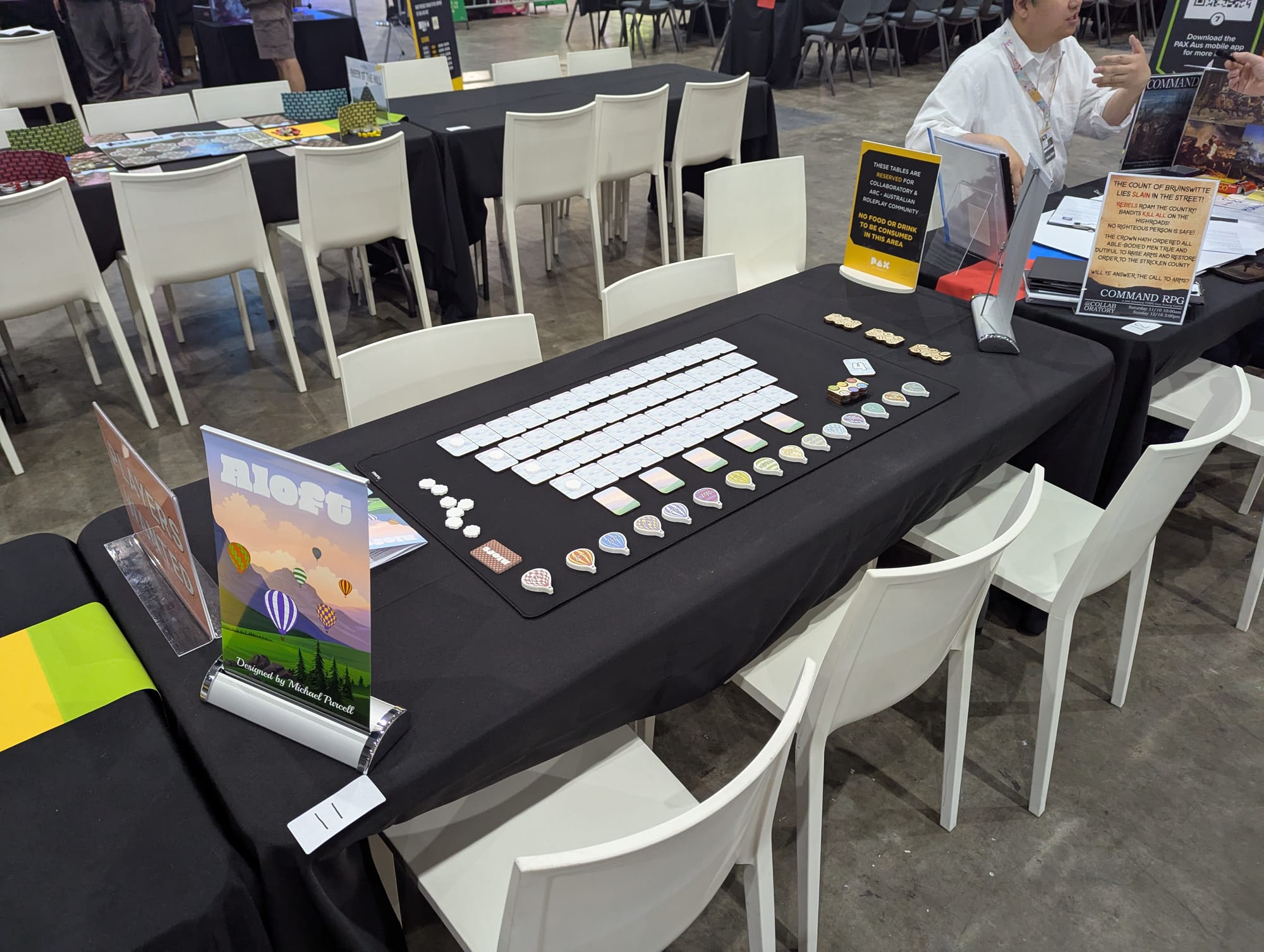 A black table at PAX with numerous tiles set out for a board game demonstration. In the foreground is an extensible sign for Aloft with a landscape of balloons.
